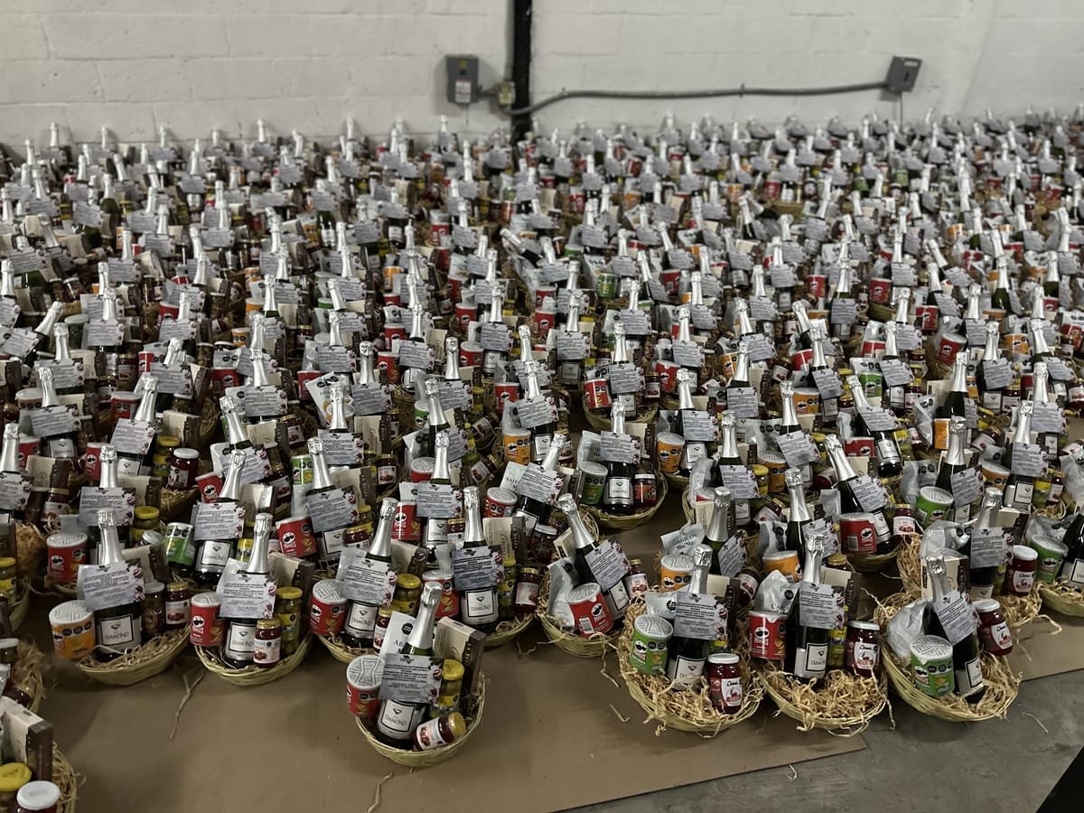 Cientos de canastas navideñas empresariales con vino espumoso, botanas y tarjeta de dedicatoria alineadas para un pedido corporativo por volumen