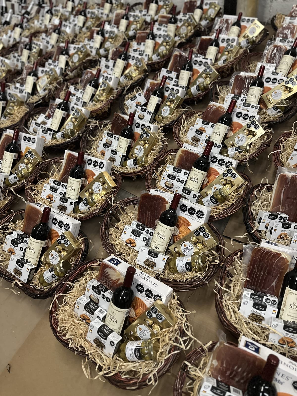 Canastas navideñas premium con vino tinto, jamón serrano, chocolates finos, aceitunas y conservas gourmet sobre paja, listas para entrega corporativa